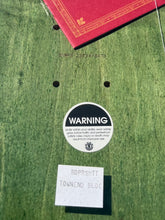 Load image into Gallery viewer, A close-up of a green wooden surface with a round black-and-white warning sticker, a small white handwritten label, and part of a red book visible at the top edge.
