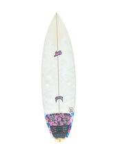 Load image into Gallery viewer, A long, narrow surfboard shown upright against a plain white background. The board is predominantly white with a small pink graphic near the top, a black logo near the center, and a colorful patterned section near the tail with pink, blue, and black tones.