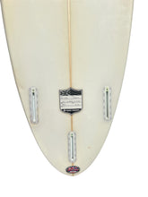 Load image into Gallery viewer, Close-up of the tail end of a cream-colored surfboard showing the three metal leash attachments, a central black and white shield logo, and a small Hawaiian logo at the bottom.