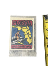 Load image into Gallery viewer, A vintage-style Florida souvenir card or matchbook cover shown next to a yellow tape measure on a plain white background. The card features a map of Florida in yellow and blue with a surfer illustration and the text 'FLORIDA' and 'THE SUNSHINE STATE'.