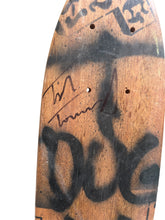 Load image into Gallery viewer, Close-up of a skateboard deck showing the underside with visible screw holes, black spray-paint graffiti markings, and a signature on a weathered wood-grain surface.