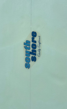 Load image into Gallery viewer, A close-up of a light blue or pale aqua textured surface with a small central logo reading 'south shore' in blue and dark blue text on a beige shape, plus smaller script text that appears to say 'Quality Softwares'.