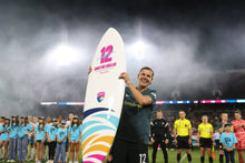 Load image into Gallery viewer, Person holding a surfboard with a number 12 on it, standing in front of a crowd at night.